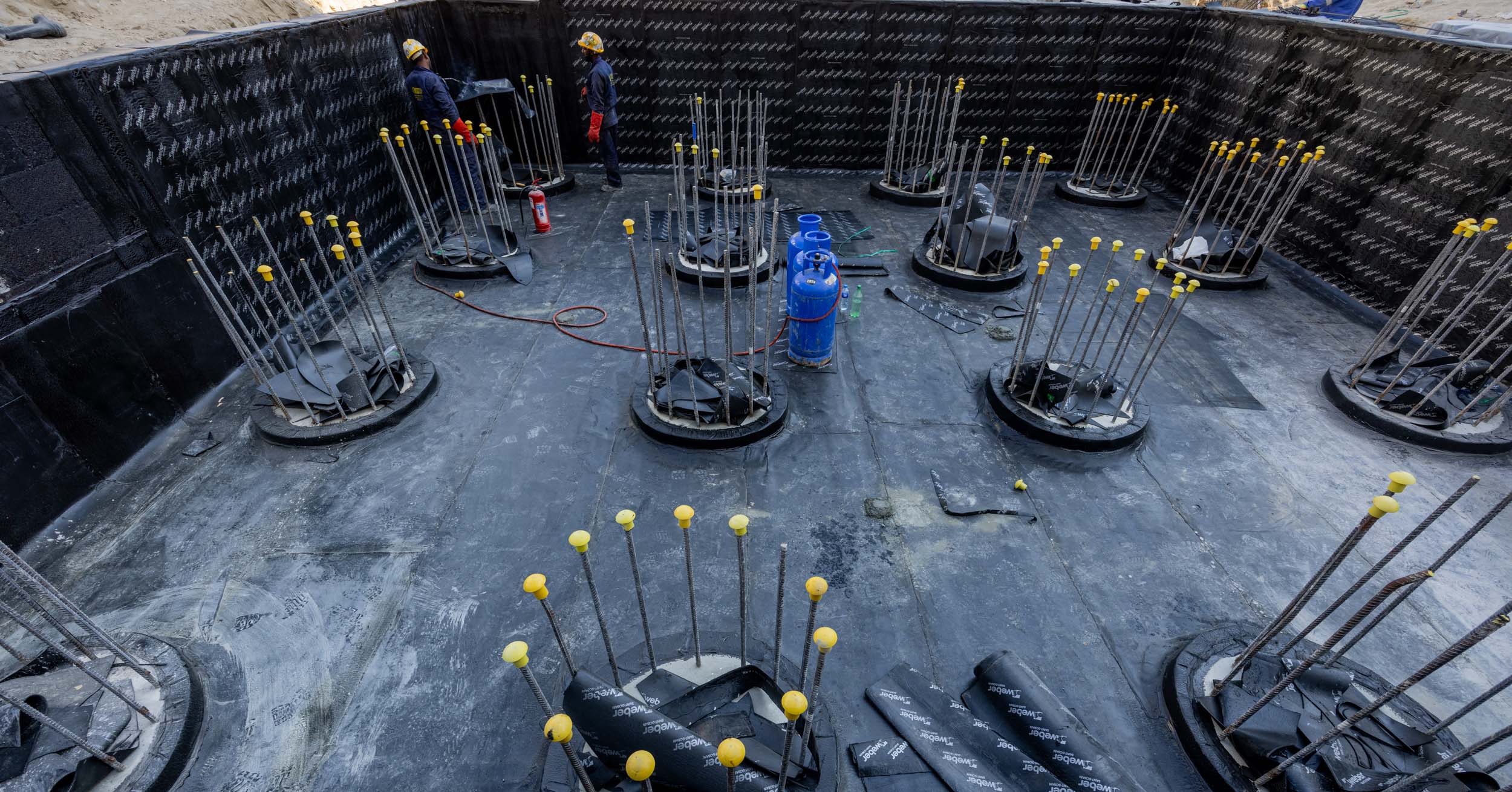 Pearl House By Imtiaz Construction site featuring circular bases with steel rebar, workers, and safety equipment in a prepared foundation area.